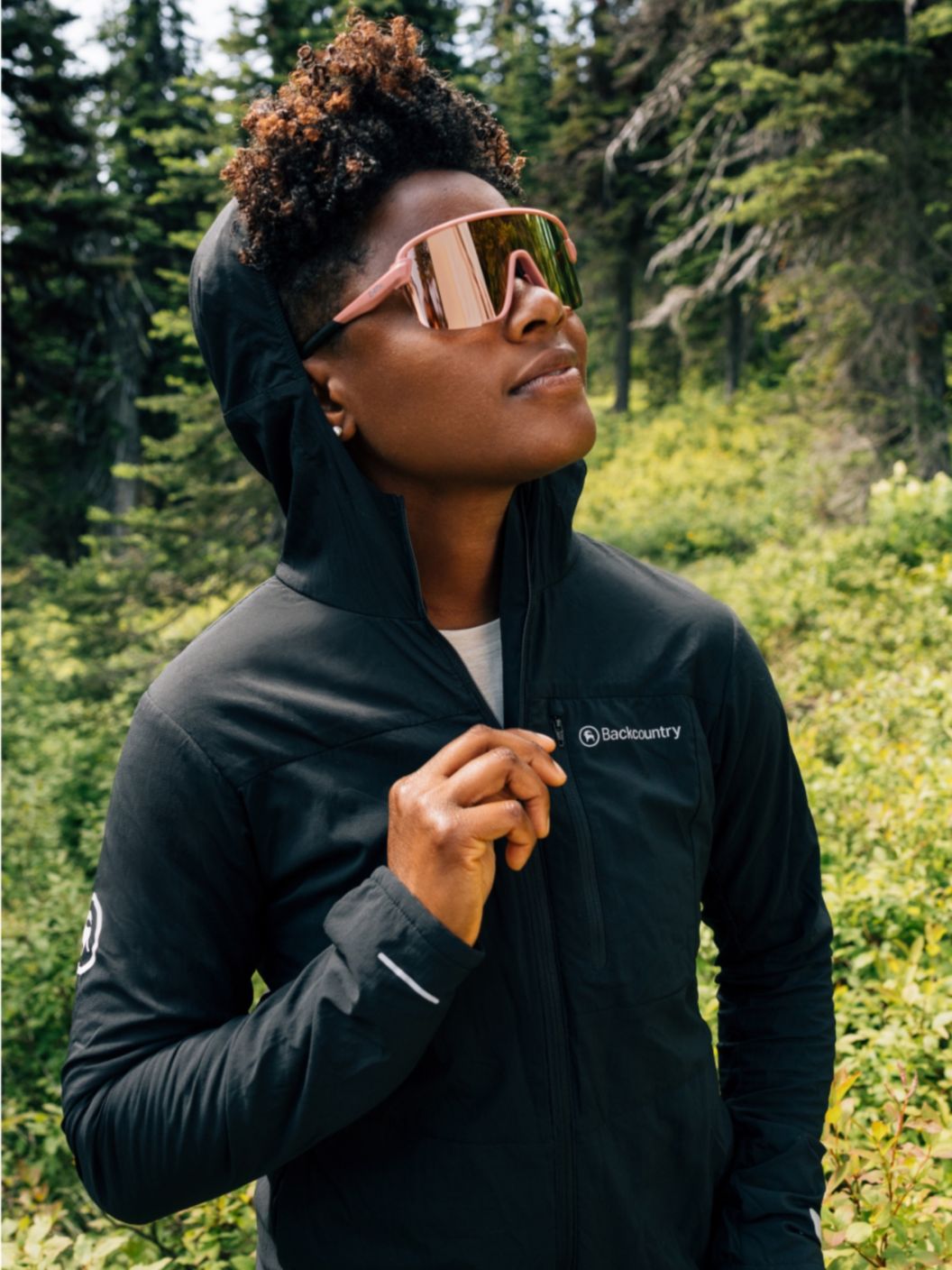 A woman in big reflective sunglasses looks up. She’s wearing a black Backcountry MTB Air Evolve jacket.